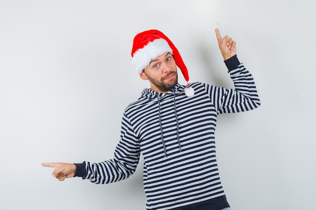 Jonge man in hoodie, kerstmuts die naar boven wijst en naar links kijkt en besluiteloos kijkt, vooraanzicht.