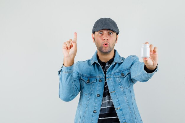 Jonge man in jas, pet met fles pillen terwijl hij naar boven wijst en verbaasd kijkt