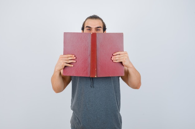 Jonge man in mouwloze hoodie met geopende boek op de mond en op zoek naar ernstige, vooraanzicht.