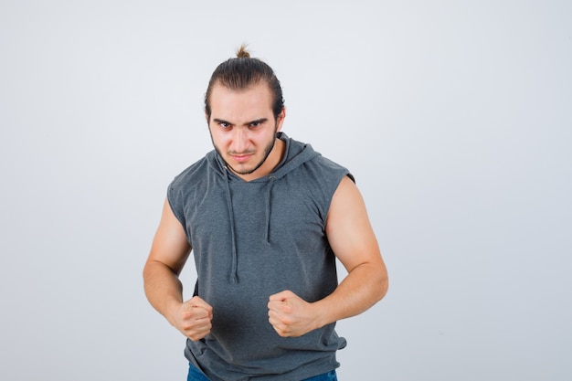 Jonge man in mouwloze hoodie staande in strijd pose en hatelijk, vooraanzicht op zoek.