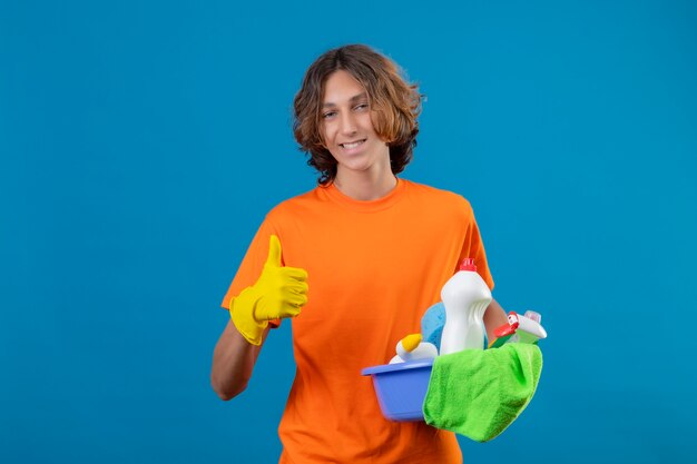 Jonge man in oranje t-shirt dragen rubberen handschoenen houden bekken met reinigingsgereedschap kijken camera glimlachend vrolijk tonen duimen omhoog staan over blauwe achtergrond