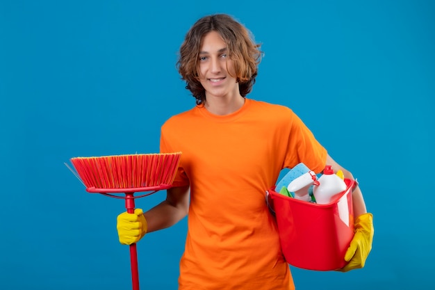 Jonge man in oranje t-shirt dragen rubberen handschoenen houden emmer met schoonmaakgereedschap en dweil camera glimlachend positief en gelukkig permanent over blauwe achtergrond kijken
