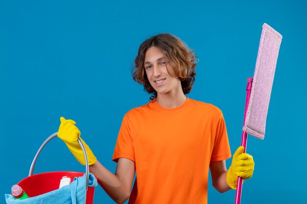 Jonge man in oranje t-shirt dragen rubberen handschoenen houden emmer met schoonmaakgereedschap en dweil camera glimlachend vrolijk blij en positief permanent over blauwe achtergrond kijken