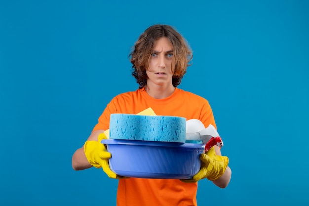 Jonge man in oranje t-shirt dragen rubberen handschoenen met bekken met reinigingsgereedschap camera kijken ontevreden met boze uitdrukking op gezicht staande over blauwe achtergrond