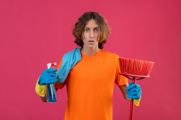 Jonge man in oranje t-shirt dragen rubberen handschoenen met dweil en schoonmaak spray camera kijken nerveus en angstig staande over roze achtergrond