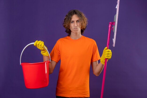 Jonge man in oranje t-shirt dragen rubberen handschoenen met emmer en dweil camera kijken met zelfverzekerde ernstige uitdrukking staande over paarse achtergrond