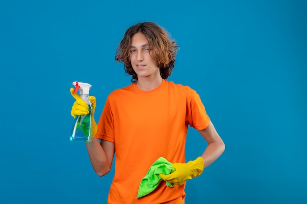 Jonge man in oranje t-shirt dragen rubberen handschoenen met reinigingsspray en tapijt camera kijken met zelfverzekerde glimlach klaar om schoon te maken staande over blauwe achtergrond