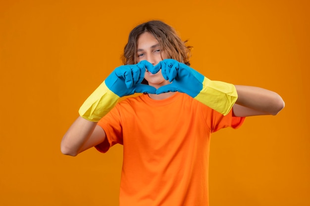 Jonge man in oranje t-shirt dragen rubberen handschoenen romantische hart gebaar kijken camera met glimlach op gezicht staan ?? staande over gele achtergrond