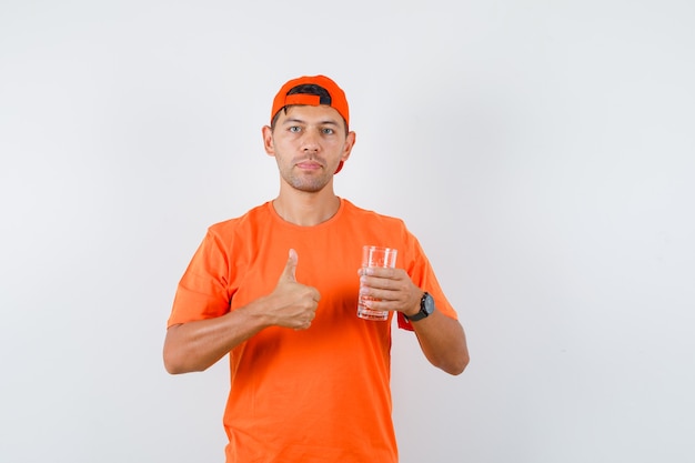 Jonge man in oranje t-shirt en pet glas water met duim omhoog te houden