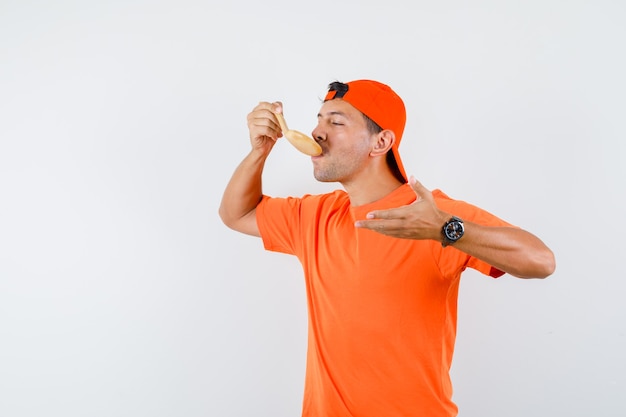 Jonge man in oranje t-shirt en pet maaltijd met houten lepel eten en hongerig kijken