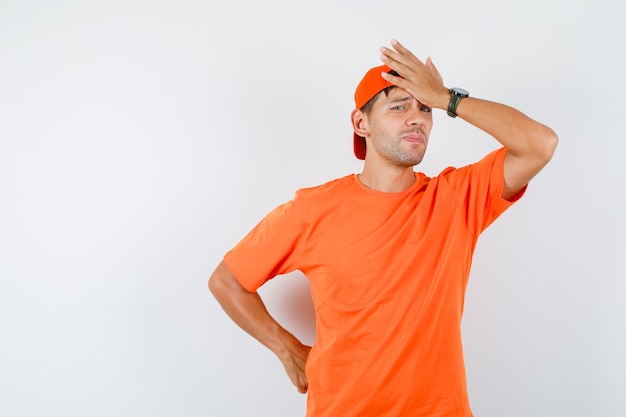 Jonge man in oranje t-shirt en pet met hand op het voorhoofd en op zoek sorry