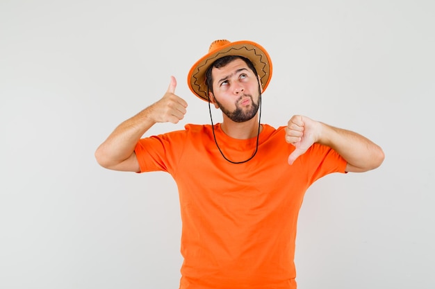 Jonge man in oranje t-shirt, hoed met duimen op en neer en aarzelend, vooraanzicht.