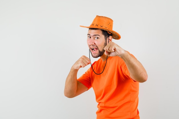 Jonge man in oranje t-shirt, hoed staat in gevechtshouding en ziet er krachtig uit, vooraanzicht.