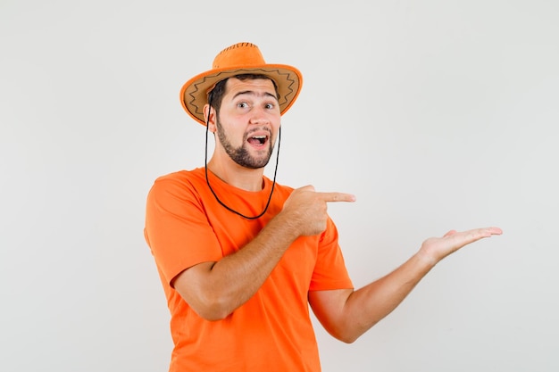 Jonge man in oranje t-shirt, hoed wijzend op palm opzij gespreid, vooraanzicht.