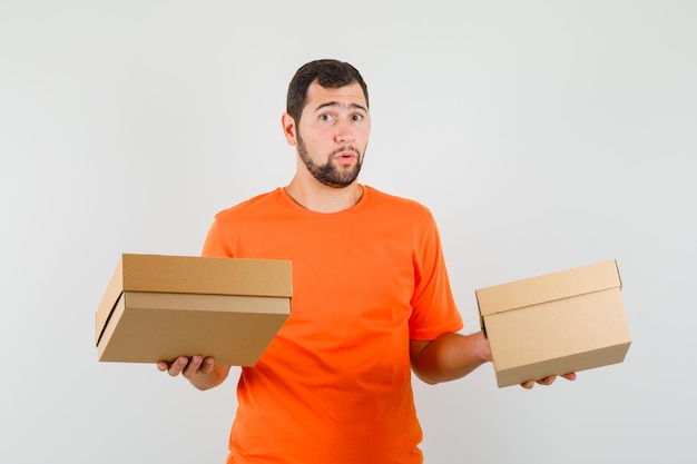 Jonge man in oranje t-shirt met kartonnen dozen en verward, vooraanzicht.
