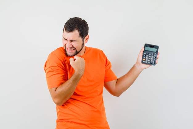 Gratis foto jonge man in oranje t-shirt met rekenmachine met winnaargebaar en zalig, vooraanzicht.