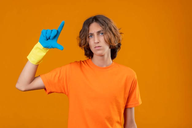 Jonge man in oranje t-shirt met rubberen handschoenen gebaren met hand weergegeven: klein formaat bord met ernstige gezicht maatregel symbool staande over gele achtergrond