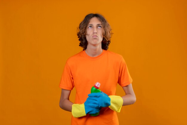 Jonge man in oranje t-shirt met rubberen handschoenen met fles schoonmaakproducten opzoeken met peinzende uitdrukking staande over gele achtergrond