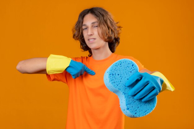 Jonge man in oranje t-shirt met rubberen handschoenen met spons wijzend naar zichzelf op zoek zelfverzekerd zelfvoldaan staande op gele achtergrond