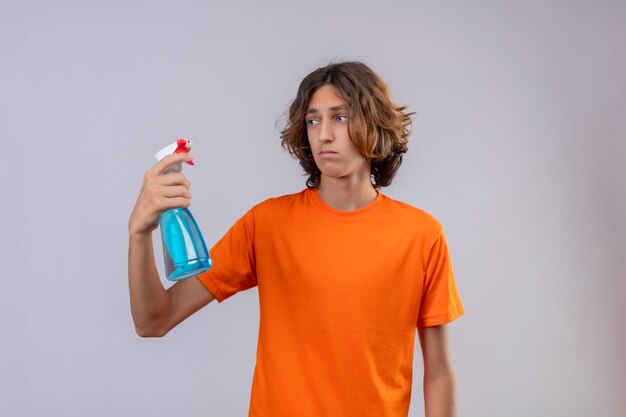 Jonge man in oranje t-shirt met schoonmaak spray te kijken met droevige uitdrukking op gezicht staande op een witte achtergrond