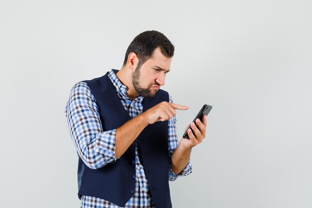 Jonge man in overhemd, vest met behulp van mobiele telefoon en op zoek naar drukke, vooraanzicht.