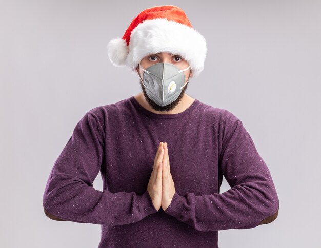 Jonge man in paarse trui en kerstmuts dragen gezicht beschermend masker hand in hand samen zoals bidden camera kijken bezorgd staande op witte achtergrond
