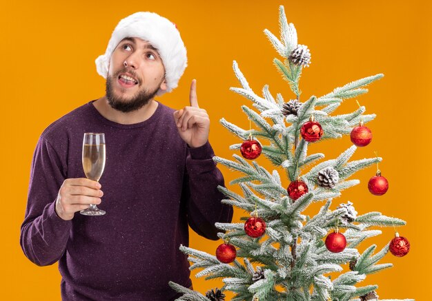 Jonge man in paarse trui en kerstmuts met glas champagne opzoeken met een glimlach op slim gezicht met wijsvinger die naast de kerstboom staat over oranje achtergrond