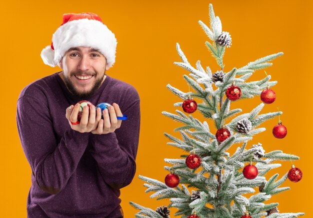Jonge man in paarse trui en kerstmuts met kerstballen blij en opgewonden lachend staande naast kerstboom over oranje achtergrond