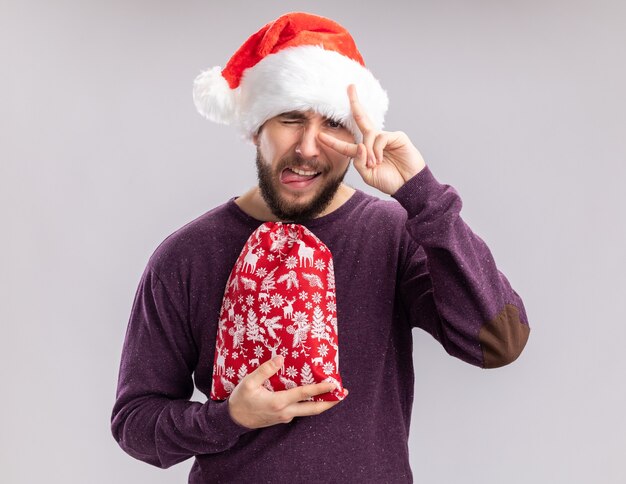 Jonge man in paarse trui en kerstmuts met rode tas met geschenken weergegeven: v-teken in de buurt van ogen tong uitsteekt staande op witte achtergrond