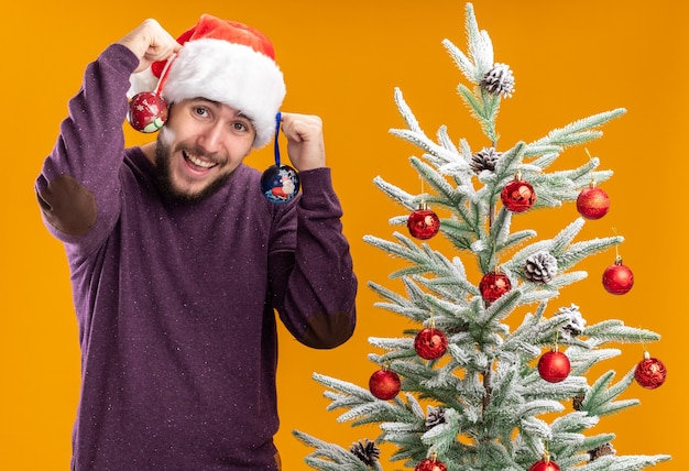 Jonge man in paarse trui en kerstmuts met speelgoed met blij gezicht lachend staande naast kerstboom over oranje muur