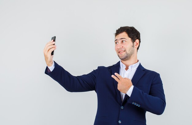 Jonge man in pak met twee vingers tijdens het nemen van selfie en op zoek vrolijk, vooraanzicht.