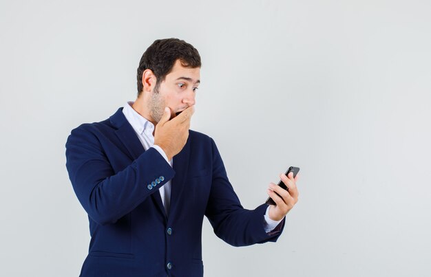 Jonge man in pak smartphone met hand op mond kijken en geschokt, vooraanzicht kijken.