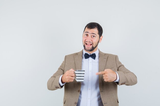 Jonge man in pak wijzend op kopje drank en op zoek optimistisch, vooraanzicht.