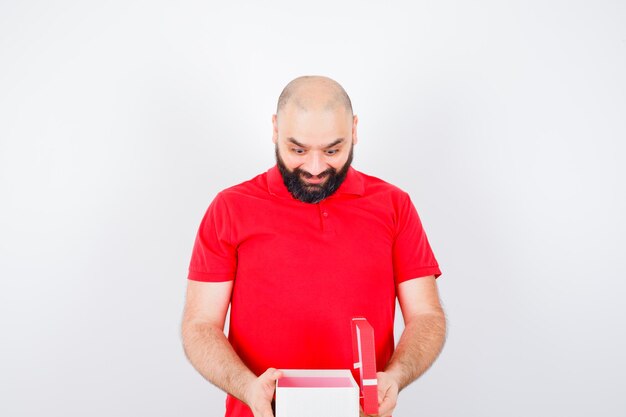 Jonge man in rood shirt die geschenkdoos opent en er gelukkig uitziet, vooraanzicht.