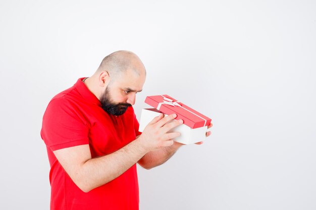 Jonge man in rood t-shirt die in geschenkdoos kijkt en zich afvraagt, vooraanzicht.