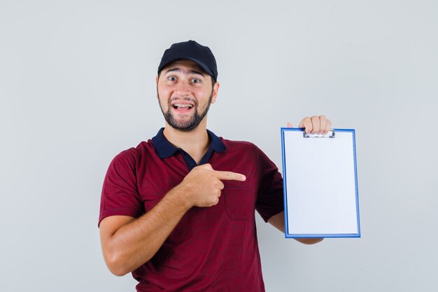 Jonge man in rood t-shirt, zwarte pet wijzend op zijn notitieboekje en kijkt tevreden, vooraanzicht.