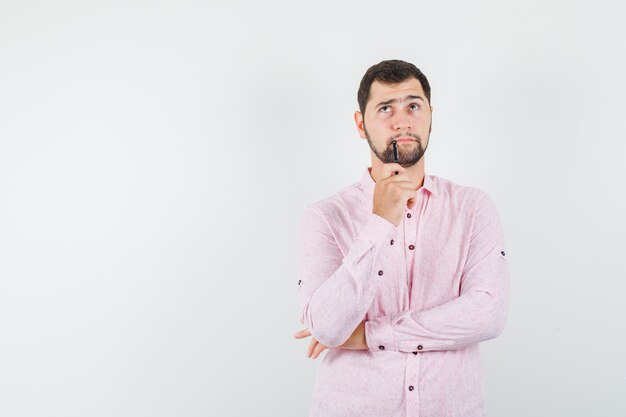 Jonge man in roze shirt met pen terwijl hij omhoog keek en aarzelend kijkt