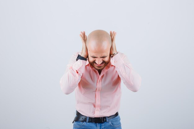 Jonge man in roze shirt, spijkerbroek hand in hand op oren terwijl hij naar voren buigt en er stressvol uitziet, vooraanzicht.