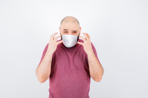 Jonge man in roze t-shirt, masker die handen op een agressieve manier opheft en er nerveus uitziet, vooraanzicht.