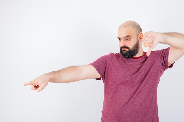 Jonge man in roze t-shirt met duim naar beneden terwijl hij wegwijst en nerveus kijkt, vooraanzicht.