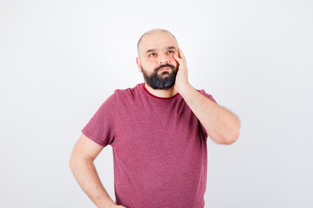 Jonge man in roze t-shirt wegkijkend terwijl hij de hand op het gezicht houdt en peinzend kijkt, vooraanzicht.