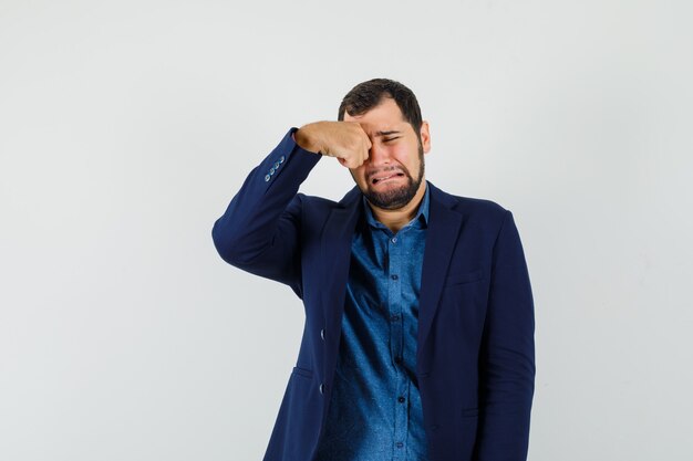 Jonge man in shirt, jasje oog wrijven terwijl huilen als een kind en beledigd op zoek, vooraanzicht.