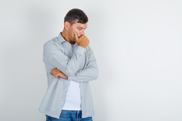 Jonge man in shirt, spijkerbroek denken met hand op kin