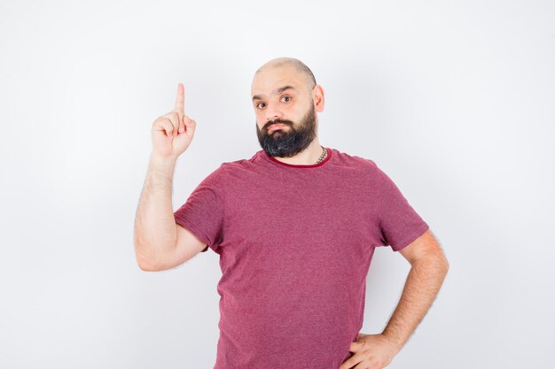 Jonge man in t-shirt die naar boven wijst en zich afvraagt, vooraanzicht.