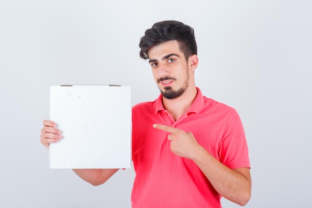 Jonge man in t-shirt die pizzadoos wijst en er vrolijk uitziet, vooraanzicht.