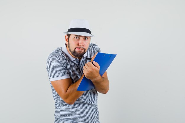 Jonge man in t-shirt, hoed die aantekeningen maakt op het klembord en er grappig uitziet, vooraanzicht.