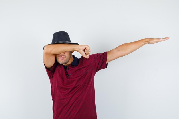 Jonge man in t-shirt, hoed die een schar bewegingsgebaar maakt en er gelukkig uitziet, vooraanzicht.