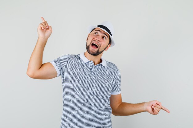 Jonge man in t-shirt, hoed die met de vingers op en neer wijst en er vrolijk uitziet, vooraanzicht.