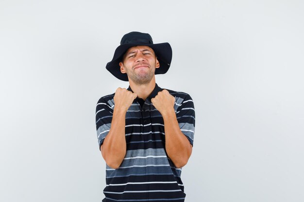 Jonge man in t-shirt, hoed die winnaargebaar toont en er gelukkig uitziet, vooraanzicht.