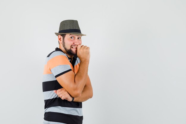 Jonge man in t-shirt, hoed staande in denken pose en op zoek vrolijk, vooraanzicht.